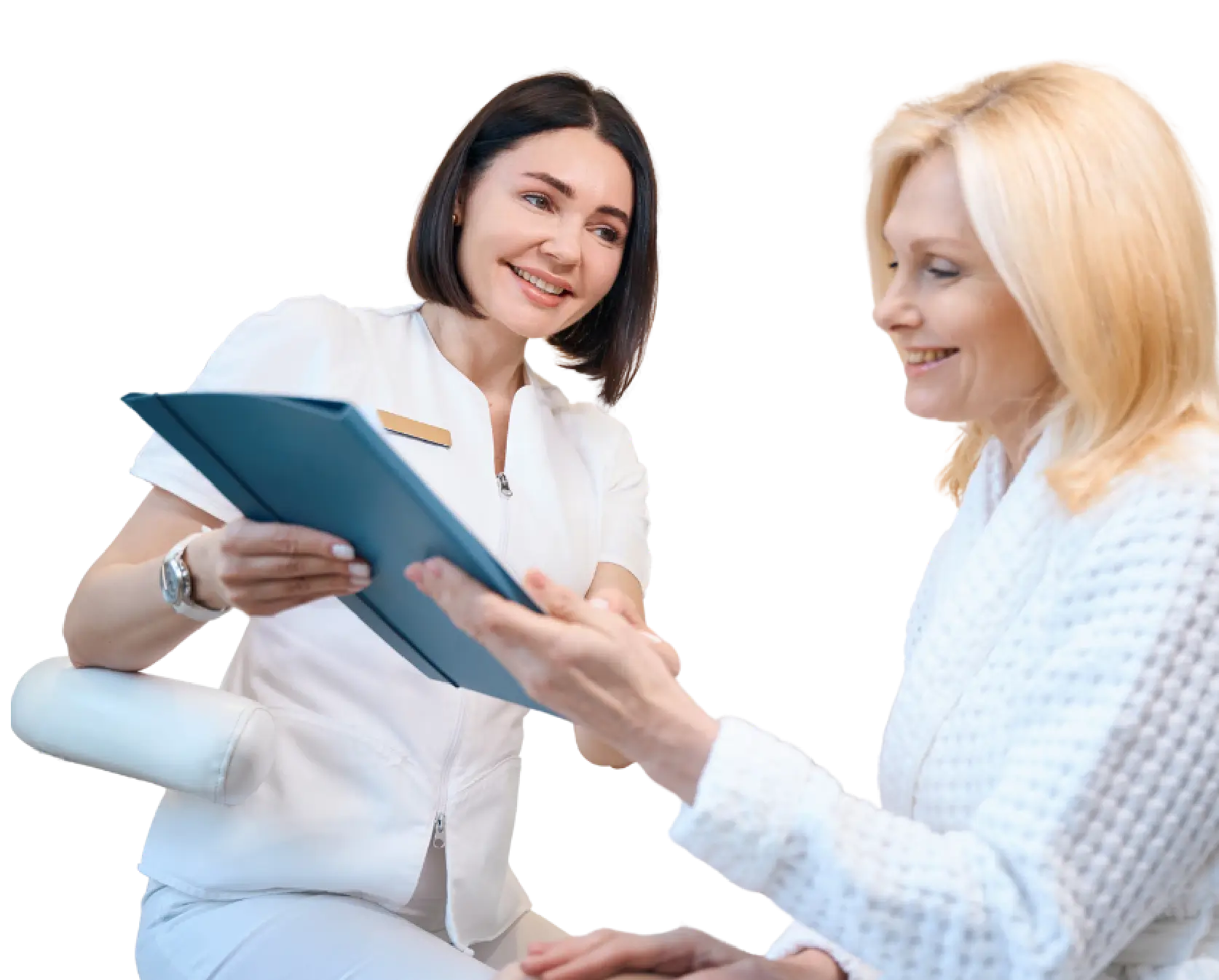A woman sits on a chair while another woman stands beside her, holding a tablet and engaging in conversation.